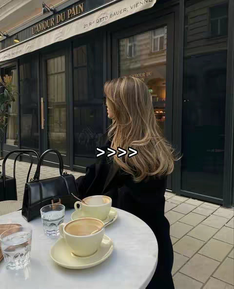 A woman sits at a cafe table with two cups of coffee, looking away from the camera. The image has a soft, warm color palette. In the background, a sign says 'L'AMOUR DU PAIN'.