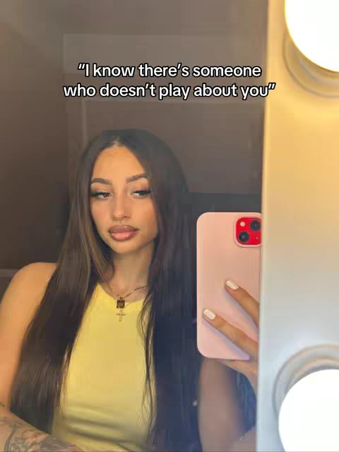 A woman with long, dark brown hair is taking a mirror selfie. She is wearing a yellow top and a necklace. A pink phone is held up to the mirror, reflecting her face and the bright lights on the side of the mirror.