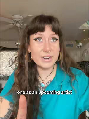 A woman with long brown hair and bangs is facing the camera. She's wearing a teal shirt, gold hoop earrings, and a necklace. The background includes a ceiling fan, a window, and some decorative elements.