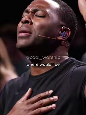 A close-up shot of a Black man in a black top, eyes closed, with a microphone in his ear, and his hand on his chest. The background is dark and blurred.