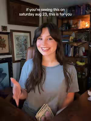 A woman with long brown hair smiles. The background includes bookshelves, artwork, and a warm, inviting ambiance.