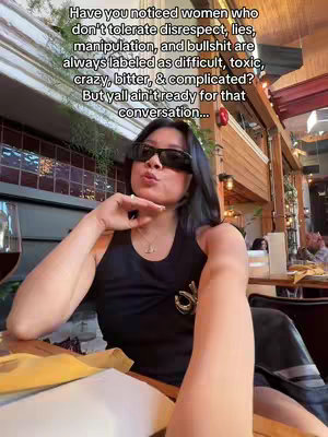 A selfie of a woman at a restaurant, wearing sunglasses and a low-cut black top, with text overlay. She has dark hair and is looking towards the camera with a slight smirk, hand near her mouth, and is seated in a booth with plants, a wine glass visible, and other diners blurred in the background.