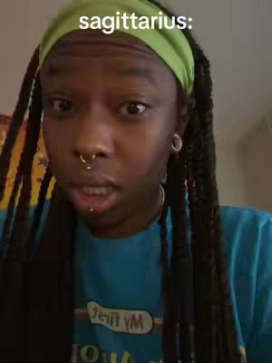 A close-up shot of a person with dark skin looking towards the camera and talking. The person has black braided hair, and is wearing a green headband and a light blue t-shirt, with a nose ring, and lip piercing. Text overlaid on the top reads "sagittarius:". The background is blurred.