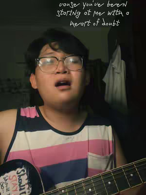 A young person with black hair wearing glasses, a striped shirt, and a guitar, singing with a visible text overlay.