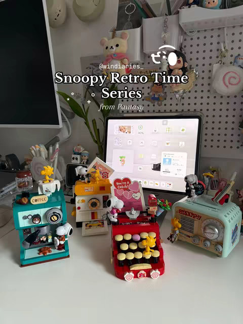 A collection of colorful Snoopy-themed Lego sets and other Snoopy figurines are displayed on a white desk. The sets are arranged to suggest a retail display, with each set on its own section of a multi-level shelf. A tablet displays an open app resembling the Lego catalog interface, partially visible. The overall composition is well-lit.