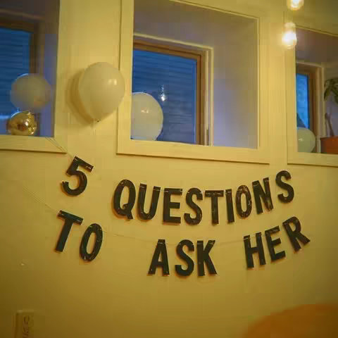 A room interior featuring a wall with several windows framing the scene. Balloons are hanging near the windows and a banner reading "5 QUESTIONS TO ASK HER" is strung across the wall. The overall lighting is warm and inviting.