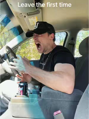 A man in a car is captured from the chest up, with the focus on his expressive face. He has a hat and is wearing a black t-shirt. The interior of the car is in soft focus and includes a can of a beverage in the console.