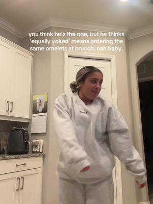 A woman is posing in a kitchen, wearing a light-colored sweatshirt and sweatpants. Text overlaid on the image reads, "you think he's the one, but he thinks 'equally yoked' means ordering the same omelets at brunch. nah baby."