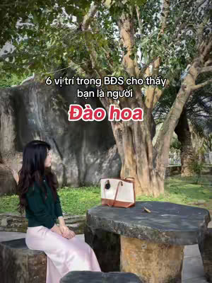 A woman sits on a stone bench with a purse on a stone table in front of a rock and with a tree in the background. Overlay text reads '6 vị trí trong BĐS cho thấy bạn là người Đào hoa'.