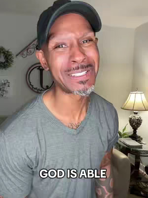 A medium shot features a smiling Black man in a gray v-neck shirt and a black cap. The background is slightly blurred, revealing a portion of a room with neutral-toned walls and decorative elements. A text overlay, 'GOD IS ABLE', is positioned at the bottom of the image.