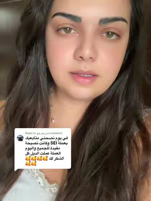 A woman with long dark hair and fair skin looks directly at the camera. She is wearing a white top, and earrings. Her hair is styled down, partially obscuring the background. Overlayed on the image is a comment with Arabic text.