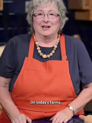 A middle-aged woman with glasses and a beaded necklace, wearing a dark blue top and an orange apron, smiles and looks directly at the camera. A text overlay at the bottom of the image reads, "On today's farms,".