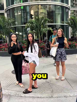 Three women stand on a sidewalk in front of a building with large glass windows. The text overlay 'you' appears in a yellow background. The woman in the middle wears a white shirt and shorts. The woman on the left wears a black top and pants. The woman on the right wears a black top and patterned skirt.