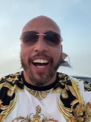 A smiling man with a bald head wearing sunglasses and a patterned shirt stands on a yacht with a light blue sky in the background.