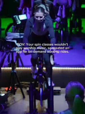 The cover image is a POV shot of a woman working out during a spin class. She is centered on the spin bike, seen from the front and slightly above with the focus on her and the equipment. A text overlay reads 'POV: Your spin classes wouldn't play worship music, so created an app for on-demand worship rides.' The background is dim with other cyclists and equipment partially visible, highlighted by green lighting. The color palette is muted with blacks, blues, and greens dominating, emphasizing the workout setting.