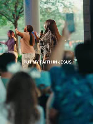 The cover image captures a live performance event, possibly a concert or church service. The overall color palette is natural with warm lighting. At the center, a woman with long brown hair is holding a microphone and appears to be singing or speaking, gesturing slightly. Behind her, there are other people, some with their hands raised, suggesting active participation. The background has a blur of trees and urban structures.