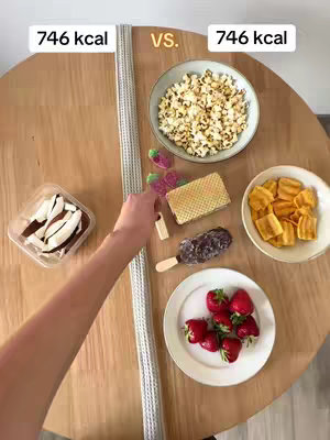 A top-down shot showcases various food items arranged on a wooden table. Two bowls, a clear container with coconut slices, and several other snacks are presented. A hand, likely of the person taking the photo, reaches in to place a piece of food next to other foods. Text overlays at the top indicate calorie counts for the items being shown.