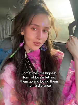 A woman with long brown hair, wearing a pink, patterned top, is sitting in a car, with the sun shining from behind. The image is a selfie-style video, where the woman is looking at the camera. Text overlay is visible at the bottom of the image.