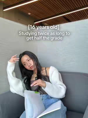 A young woman with long black hair is seated on a gray sofa, holding a book or papers. She's dressed in a white sweater and a tank top with blue jeans. Text overlays state her age and the video's theme.