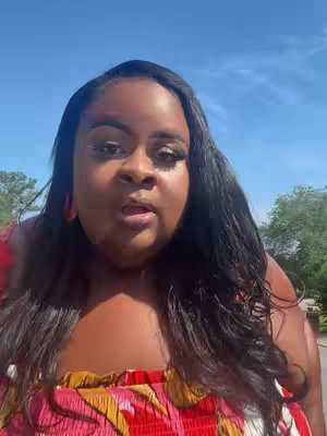 The cover image is a selfie-style shot of a woman facing the camera, framed from the chest up. The background is a sunny outdoor scene with a blue sky and glimpses of green trees and foliage, suggesting a pleasant day. The woman has dark hair and is wearing a red and patterned top. Her expression is neutral, with her mouth slightly open as though she is speaking.