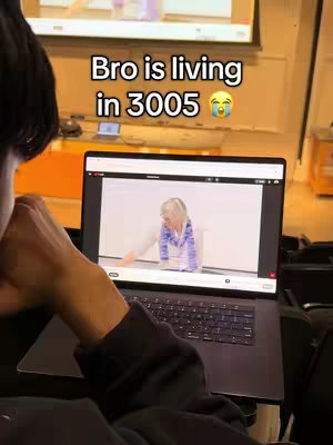 The image shows a person holding a laptop with a video playing on its screen. Above the laptop screen, another screen is visible, likely a projector or a larger display, showing the same video. Overlayed text appears on the laptop screen. The background appears to be a lecture hall or a similar academic setting, with warm lighting and an orange surface visible. The focus is on the laptop and the interaction with the content displayed.