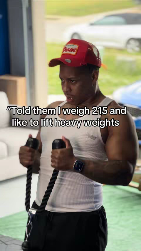 A man in a red cap and a white tank top is lifting weights with black cables. The background is a garage or a gym setting.