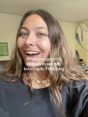A woman with brown hair and green eyes is the main focus, smiling broadly with her mouth open, seemingly surprised or excited. Gold jewelry can be seen around her neck. The background is a softly lit interior with a framed image on the wall and a partial view of what looks like a surfboard or decorative item to the right.