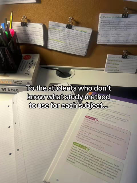 The image shows a desk with study materials. A book, notebooks, and pen holders are visible. On a board behind the materials, there are index cards clipped, with text written on them. The scene is lit with natural light.