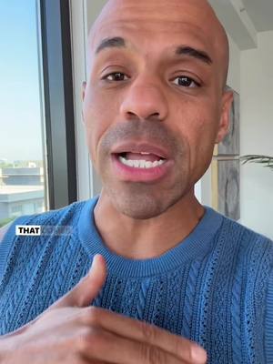 A bald man with brown skin and a light beard is looking at the camera and talking indoors. He is wearing a blue sweater and making a hand gesture. The background is windowed, with a glimpse of a cityscape in the distance.