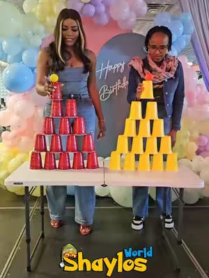 Two women are shown standing behind white folding tables, each with a stack of cups. The backdrop has a circle saying Happy Birthday. One stack is red and one stack is yellow.