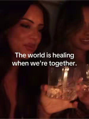 The image shows two women close together, likely in a restaurant or similar dimly lit space, possibly a bar. One is holding a glass of a golden liquid, catching the light. The text overlay conveys a message of togetherness.