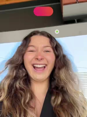 A young woman with long, wavy brown hair is captured in a close-up shot, her face expressing extreme happiness and excitement. Her mouth is wide open in a laugh, revealing her teeth, and she seems to be in mid-speech or reaction. The background appears blurred, possibly an interior space with overhead architectural elements and light blue tones in the background.