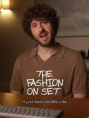 A medium shot shows a man with curly brown hair wearing a brown shirt, looking directly at the camera. The text overlay includes the video hook, “THE FASHION ON SET”. Below the man, the words “It just feels like 90s vibe”are present. The background appears to be inside a room with warm lighting and a glimpse of a desk, further suggesting a behind-the-scenes or casual setting.