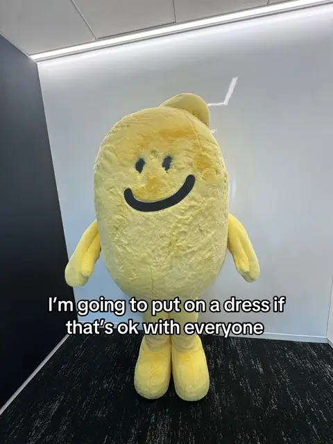 A full-body shot features a person wearing a yellow, egg-shaped costume with a smiling face. The costume has arms and legs. The person is standing against a background of a white wall, a black and gray patterned floor, and a dark-colored wall.