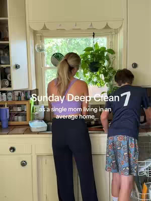 A person viewed from the waist up, facing away from the camera, is standing in front of a kitchen sink with a young person. The person is wearing a light purple top with black pants. The young person is wearing a dark shirt with a number 7 on the back and blue patterned shorts. The kitchen has cream-colored cabinets and a window with greenery visible through it.