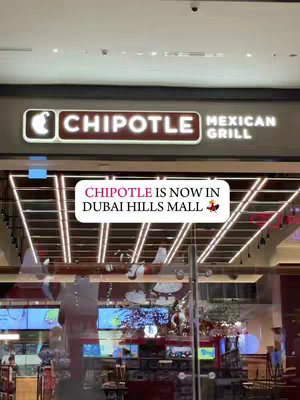The cover image is taken from inside a mall, showcasing the entrance of a Chipotle restaurant. The sign is prominently displayed with the restaurant's name and logo in a brown and white color scheme. Below the sign, there is a text overlay in a white rectangle that reads 'CHIPOTLE IS NOW IN DUBAI HILLS MALL', accompanied by a taco emoji. The background reveals the interior of the restaurant, which includes customers, tables, the ordering counter, and the menu on the screen. The lighting is bright, with a combination of natural light and spotlights.