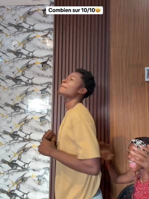 A man in a yellow shirt stands with his head tilted back, eyes closed, and hands near his chest. Another person's hand is reaching over to cover the face of another person. The text overlay reads 'Combien sur 10/10'. The backdrop includes a marble patterned wall.
