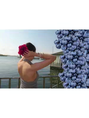 A woman standing on a deck with her back to the camera, arms raised to hold her hair, with a large red flower in her hair. To the right is a column of blueberries. The background features a body of water, buildings, and a bright sky.