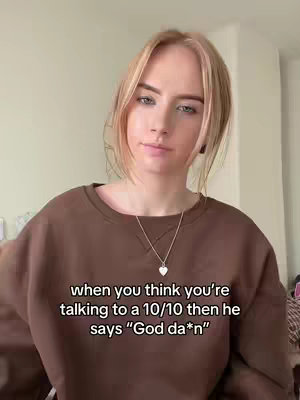 A young woman with blonde hair and blue eyes is centered in the frame, wearing a brown sweatshirt and a silver heart necklace. She has a slight smirk on her face. Text overlay is at the bottom of the image. The background is a plain, light-colored wall.