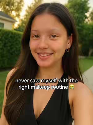 A young woman with long dark hair smiles at the camera. She's in front of some green foliage, and a house is in the background. Text overlayed on the video reads "never saw myself with the right makeup routine"