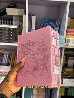 A person with dark skin holds a pink Bible open towards the viewer, showcasing the tabs and floral design on the cover. The book sits amidst a white-shelved bookcase filled with various books. The scene is well-lit, and the focus is sharp on the Bible.