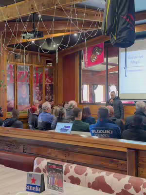 A group of people seated in a wooden-paneled room, with one person standing and speaking into a microphone. A projector screen is visible with text related to the 'Executive Mayor.' The room has string lights and a flag hanging. Foreground has tables with the Heineken logo and other promotional materials.