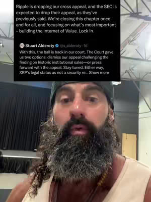 A selfie-style video frame featuring a man with a thick, graying beard and a black cap. The man is centered, filling a good portion of the frame. Above him, overlaid on the image, is a screenshot of a tweet from Stuart Alderoty discussing Ripple dropping an appeal, with text from the tweet visible. The background is out of focus, suggesting an indoor setting with possible gym equipment or studio lights, adding a sense of urgency.