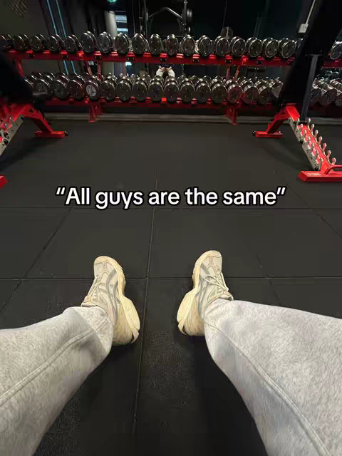 The image shows a first-person perspective of a person's legs and feet with grey sweatpants and beige sneakers in a gym. In the blurred background, there are rows of dumbbells.