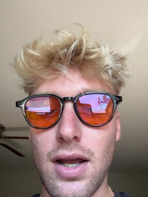 A close-up shot of a young man wearing dark-framed sunglasses with orange lenses. The man has light-colored hair and a light complexion. The background is blurred and appears to be an interior space.
