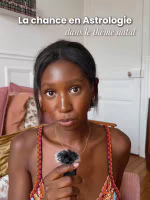 The cover image features a woman holding a small microphone in front of her mouth, gazing directly at the camera. She is framed from the chest up. The background is a soft, blurred interior with a glimpse of a bed and a decorative framed artwork, suggesting a home setting. Above her head, superimposed text reads 'La chance en Astrologie dans le thème natal'.