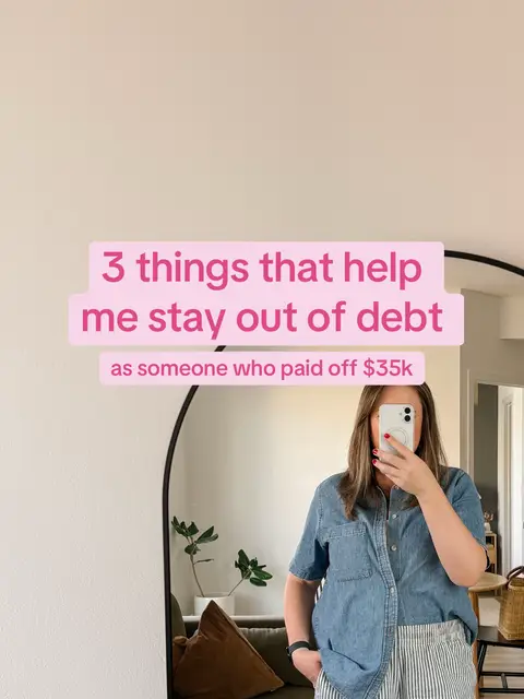 A person is seen in front of a large, arched mirror. They are taking a selfie with their phone, obscuring their face. The person is wearing a denim shirt and striped pants. Behind them, the room includes a sofa, a small plant, and a wicker basket. Text overlay in pink rectangles states: "3 things that help me stay out of debt as someone who paid off $35k."