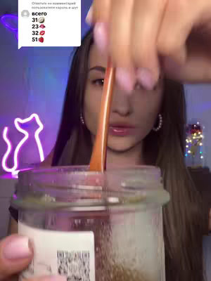 A woman is seen face-on, stirring a clear liquid inside a glass jar with a light orange spoon. Her gaze is slightly downward. In the background, a neon cat silhouette is visible on one side, while the other has fairy lights.