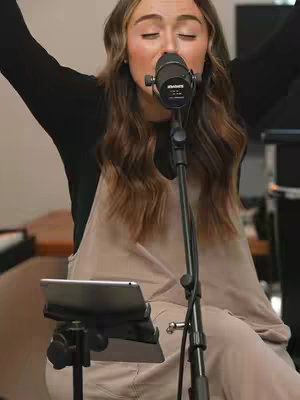 A woman with long brown hair stands centered, holding a microphone. She has her eyes closed and arms raised, set against a blurred background with various equipment. She wears a black top and a beige jumpsuit.