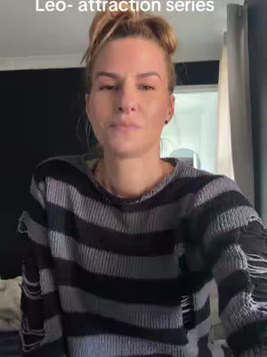 A woman with light skin and blonde hair in a bun is centered in the frame, looking towards the camera, wearing a black and grey striped sweater. The background includes a black wall and a gray curtain. The text overlay displays the title of the series.